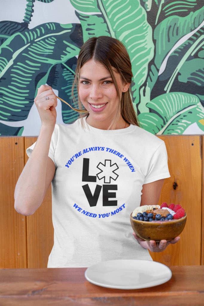 Woman in a white EMS Hero Hues t-shirt with star of life and "LOVE" graphic, eating a healthy fruit bowl.
